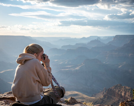Blond young girl with sweatshirt sitting on the rock of the precipice of the grand canyon, she is taking photos of the beautiful landscape
