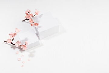Wedding gentle scene mockup - two white square podiums, twig of pink cherry flowers, petals in sunlight on white background, top view, template for presentation cosmetic products, goods, branding.
