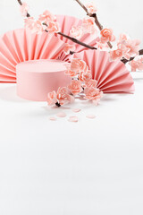 Spring stage mockup with pink cylinder podium for presentation cosmetic products, goods, asian fans, branch of pink sakura flowers on white table in traditional japanese style, copy space, vertical.