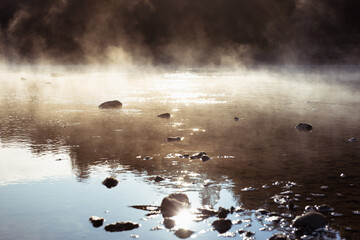 Fluffy white fog above river water in early morning in golden sunbeams with ripple sun glare,...