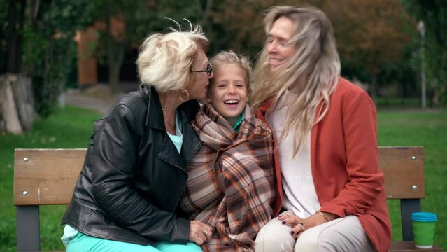Loving mother and grandmother kissing girl sitting on bench smiling looking at camera. Happy Caucasian kid posing outdoors with family in windy park