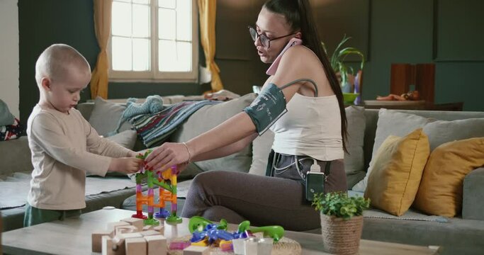 Young woman wearing holter monitor device playing with her little son at home while talking on the phone