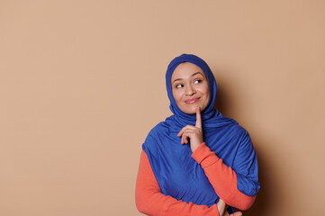 Pleasant positive Arab Muslim woman, dressed in elegant orange dress and blue hijab, putting her finger on chin, reasoning over problems, thinking, isolated on beige background. People. Lifestyle.