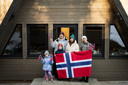 Portrait Of Family With Kids Outside Cabin House Holding Norway Flags. Scandinavian Culture, Norwegian People.