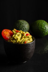 Top view of black bowl with guacamole on glossy black background with avocados, selective focus, vertical, with copy space
