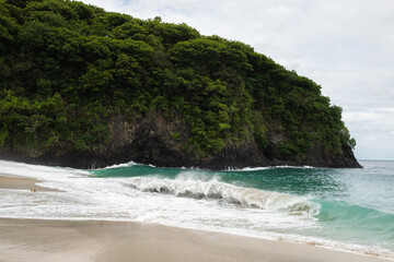 Tropical coastal landscape with green cliff, turquoise ocean waves and sandy beach on a cloudy day.