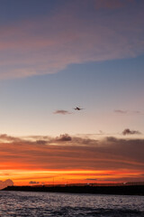 Commercial airplane landing over ocean at sunset with vibrant sky, scenic aviation travel photography.