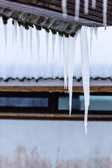 Vertical photo of icicle at old asbestos roof, background old brown narrow windows. Isolation problems in house. Icy icicle hanging and melting down of old mossy green and brown roof