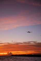 Commercial airplane landing over ocean at sunset with vibrant sky, scenic aviation travel photography.
