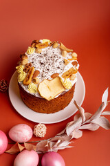 Beautiful Easter card with traditional Easter cake decorated with white sugar glaze, nuts and dried fruits on white round plate and pink painted Easter eggs from above on bright orange background. 
