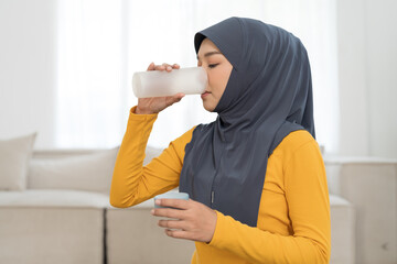 Asian young Muslim woman wearing hijab drinking water at home