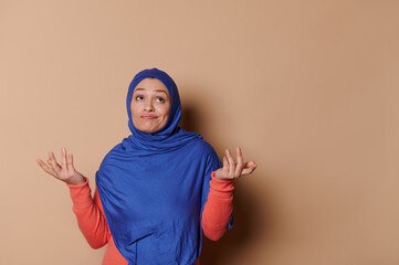 Thoughtful Arabic Muslim woman in a blue hijab, looking up with uncertainty, holding her hands palms up, standing isolated on a beige background. People. Concepts. Lifestyle