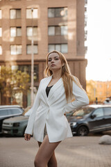 Fototapeta premium Blond stylish woman at oversized white jacket posing at the city background