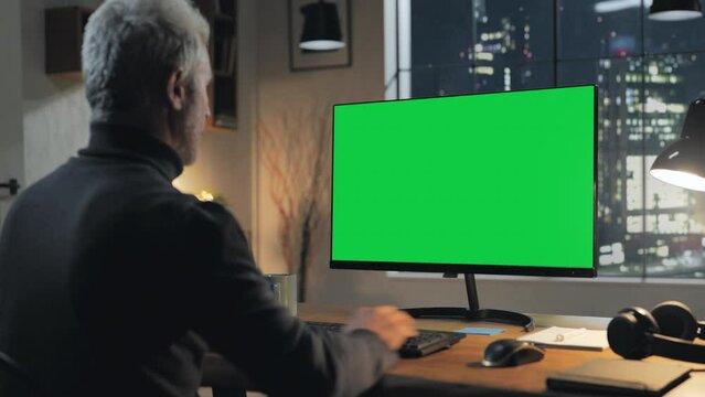 Back view of man using computer green screen in home office at night modern city view in the background,chroma key pc monitor