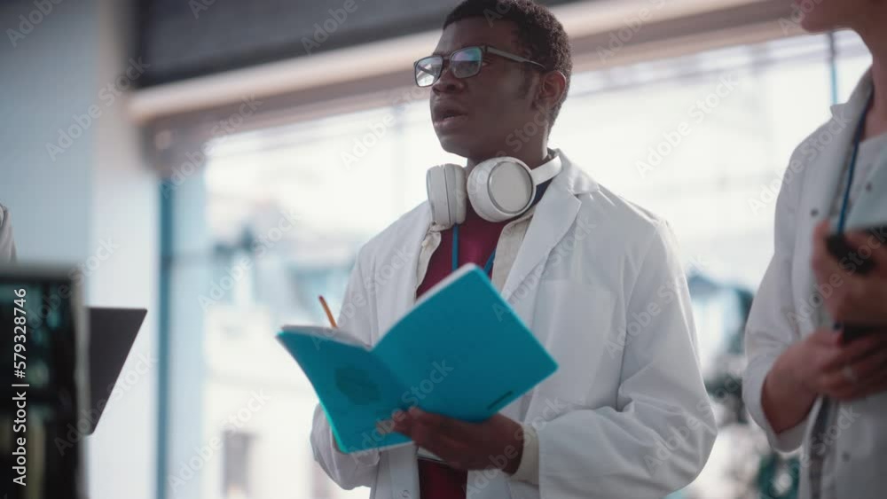 Poster Portrait of a Young Black Male Engineering Student Asking Questions and Taking Notes in a Laboratory. Lab Assistant in a Meeting Showing Initiative and Giving Ideas, Brainstorming with his Team