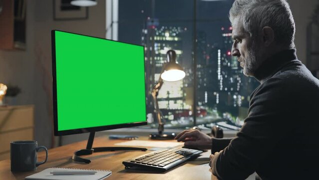 mature male at the desk using computer green screen at night,modern home office with window city view in the background,chroma key monitor