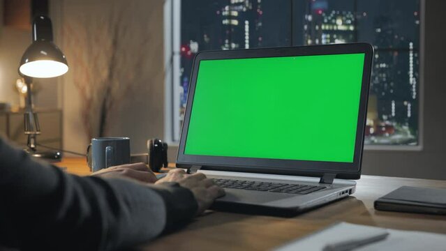 hands typing laptop computer with green screen at night window modern home office with window city view in the background,chroma key notebook monitor