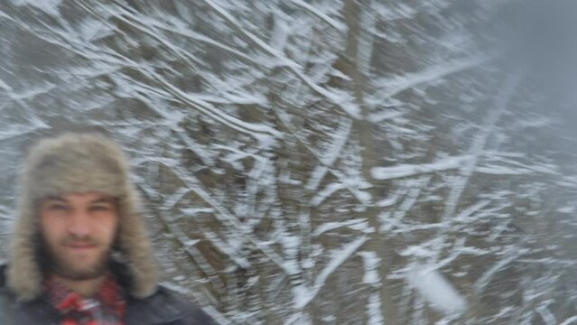 Young Caucasian man on walk outside in winter wears fluffy hat with earflaps and red plaid shirt. Portrait close-up front view. The guy throws snowball and gets right into camera lens.