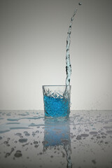 Vertical shot of water pouring into crystal glass with droplets around, on a white shiny table
