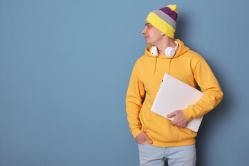 Portrait of cute positive man wearing beanie hat and casual yellow hoodie standing isolated over blue background, looking away at copy space, holding notebook, advertisement area