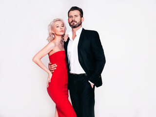 Beautiful fashion woman and her handsome boyfriend in black elegant suit. Sexy blond model in red evening dress. Fashionable couple posing in studio. Brutal man and his female. Isolated on white