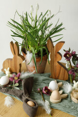 Happy Easter! Stylish wooden bunny, spring flowers, natural eggs and napkin bunny ears on rustic table in room. Easter still life. Festive arrangement and decor in farmhouse