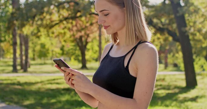 Young woman looks at running progress on phone app. Smiling sportswoman excited to see perfect results of running in autumn city park