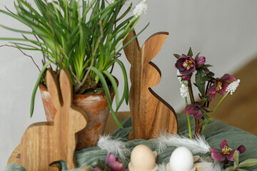 Happy Easter! Stylish wooden bunny, spring flowers, natural eggs and feathers on rustic table in room. Easter still life. Festive arrangement and decor in farmhouse