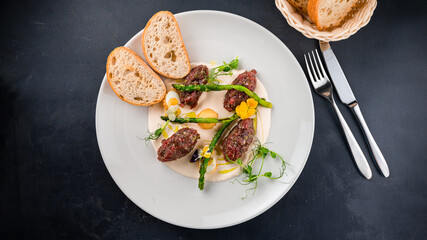 Appetizer of beef tartare with with asparagus, herbs, sauce and slices of bread.