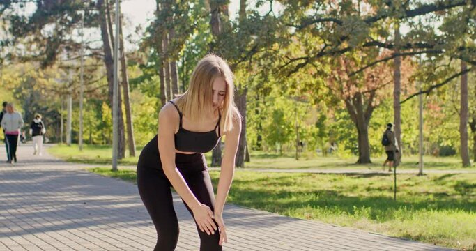 Short-haired woman stops running having leg ache in city park. Young sportswoman tries to stop knee pain and decides to take break from jogging