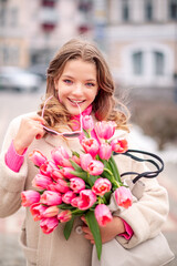 Beautiful young woman with spring tulips flowers bouquet at city street. Happy girl smiling and holding pink tulip flowers outdoors. Spring portrait of pretty female in park.