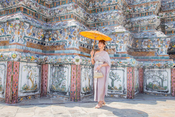Young woman wearing Thai dress with accessories stands holding antique umbrella and bag at Wat Arun Ratchawararam It is a popular destination for tourists around the world. Bangkok, Thailand