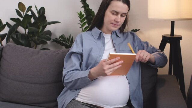 Dreamful cute smiling young pregnant woman sitting on cozy sofa and takes notes in notebook, makes list of necessary things for pregnancy, childbirth. Pretty happy mother waiting of baby at home