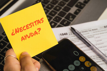 Hands holding a help text in spanish on sticky notes with 1040 tax forms , United States tax form, a laptop on a desk near a phone with a calculator app. US individual income tax return. 