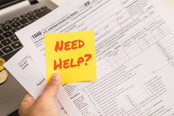 Hands holding a help text in spanish on sticky notes with 1040 tax forms , United States tax form, a laptop on a desk near some bitcoins. US individual income tax return. 