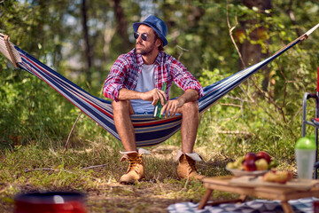 Young handsome hipster male sitting in hammock holding beer. Picnic in forest. Holiday, leisure, fun, lifestyle concept.