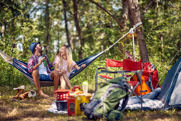 Young cheerful hipster couple having small talk and sitting in hammock, enjoying camping in nature. Picnic in forest. Holiday, leisure, fun, lifestyle concept.