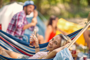 Friends having picnic in nature, outdoors. Young woman laying in hammock holding beer. Holiday,...