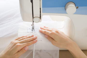 Young adult woman hands working with sewing machine and sewing edge of white day curtains. Closeup. Point of view shot. Top down view.