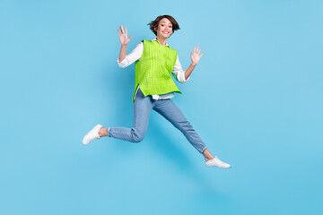 Fototapeta na wymiar Full size photo of satisfied adorable girl with brown hairdo wear green vest jeans flying jumping isolated on blue color background