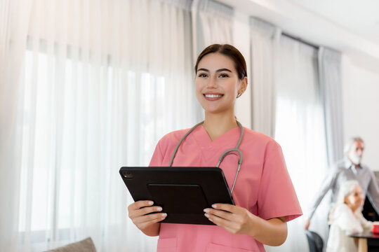 Young processional Caucasian Geriatric doctor in pink scrubs with stethoscope holding digital tablet. Physician in hospital nursing or wellbeing county. Happy caregiver taking care of elderly people.
