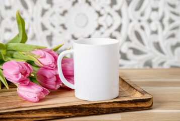Fototapeta na wymiar Selective focus on single one white mug mock up. Cup on home table with decorative spring flowers on background and bohemian style wood panel. Cozy seasonal products advertisement background.
