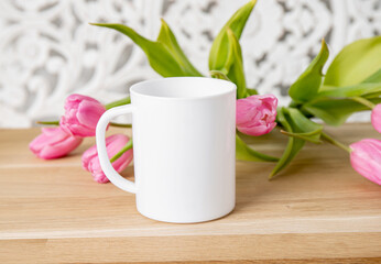 Selective focus on single one white mug mock up. Cup on home table with decorative spring flowers on background and bohemian style wood panel. Cozy seasonal products advertisement background.