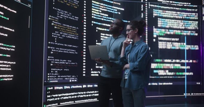 Portrait Two Diverse Developpers Using Laptop Computer, Discussing Lines of Code that Appear on Big Screens Surrounding Them. Male and Female Programmers Creating a Software Together, Fixing Bugs