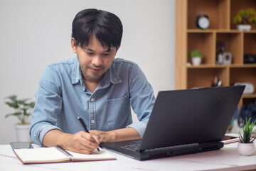 A businessman works at the front of the laptop with a note in the notebook.