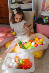 A little girl playing with colored rice and Easter eggs in sensory bin. Easter sensory bin with bright rice and eggs, bunny, carrot. Sensory play and holidays activity for kids. Happy Easter concept.