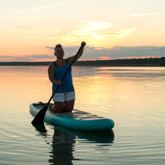 A woman with a mohawk in shorts on a sapboard with an oar against the backdrop of a bright sunset sky swims in the lake in the evening.