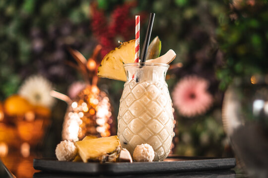 Pina Colada in dekorativem Cocktail Glas mit Trinkhalm, Ananas und Dekoration auf schwarzem Holzbrett