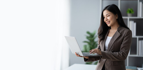 Beautiful Asian businesswoman using laptop computer while standing in the modern office room.