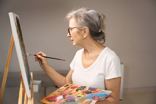 Gray-haired lady in casual clothes stands in front of easel